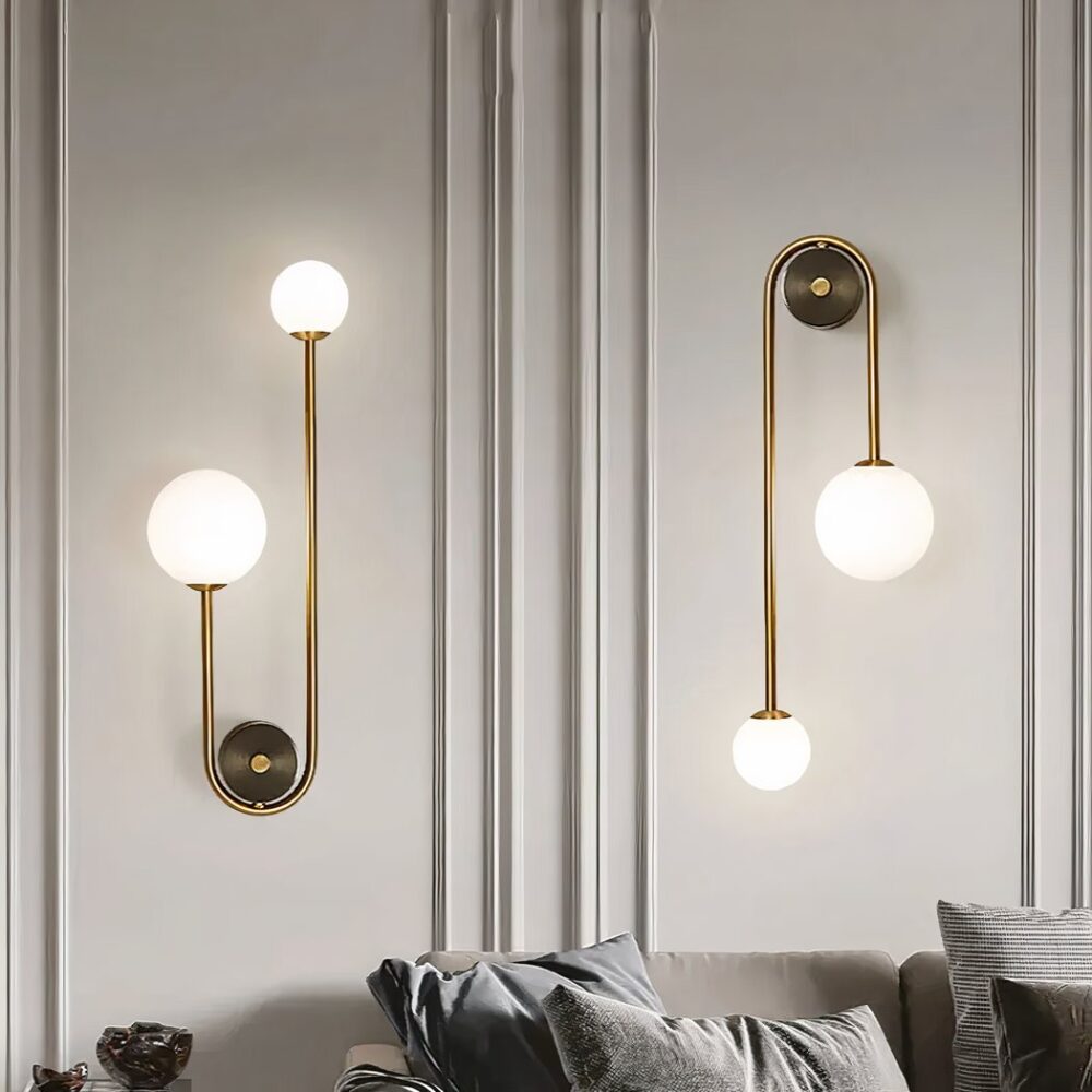 Pared gris sobre la que se fijan 2 apliques, soporte redondo dorado con una varilla dorada que va a izquierda y derecha, uno más corto que el otro. El más pequeño tiene una bola de luz grande y el más largo una pequeña. El aplique de la izquierda está fijado con las bolas hacia arriba, el de la derecha con las bolas hacia abajo. Debajo de los apliques hay un sofá gris con cojines a juego.