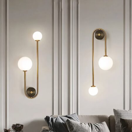 Pared gris sobre la que se fijan 2 apliques, soporte redondo dorado con una varilla dorada que va a izquierda y derecha, uno más corto que el otro. El más pequeño tiene una bola de luz grande y el más largo una pequeña. El aplique de la izquierda está fijado con las bolas hacia arriba, el de la derecha con las bolas hacia abajo. Debajo de los apliques hay un sofá gris con cojines a juego.