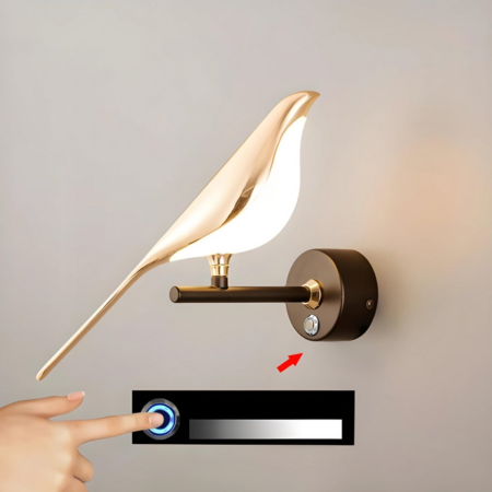 Aplique dorado de urraca con interruptor táctil, colgado en una pared beige. En la parte inferior de la foto, una mano muestra visualmente el uso del interruptor