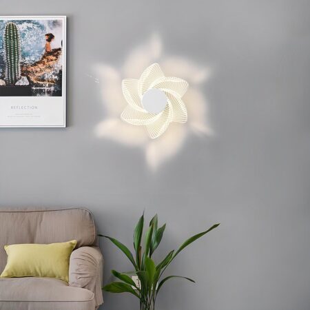 Aplique en forma de flor iluminado sobre una pared gris. A la izquierda, un cuadro y debajo un sofá beige con un cojín amarillo y a su derecha una planta verde.