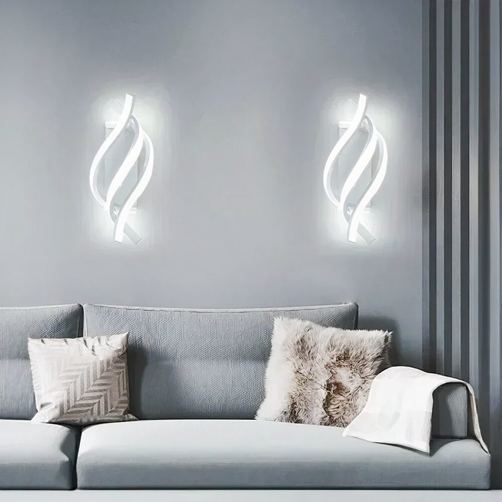 2 apliques en espiral blancos con 3 filamentos iluminados con luz fría. Montados en una pared gris, cortina gris oscuro a la derecha. Un sofá gris bajo los apliques con 2 cojines: uno blanco con un motivo dorado y otro de piel, una tela en el reposabrazos del sofá.