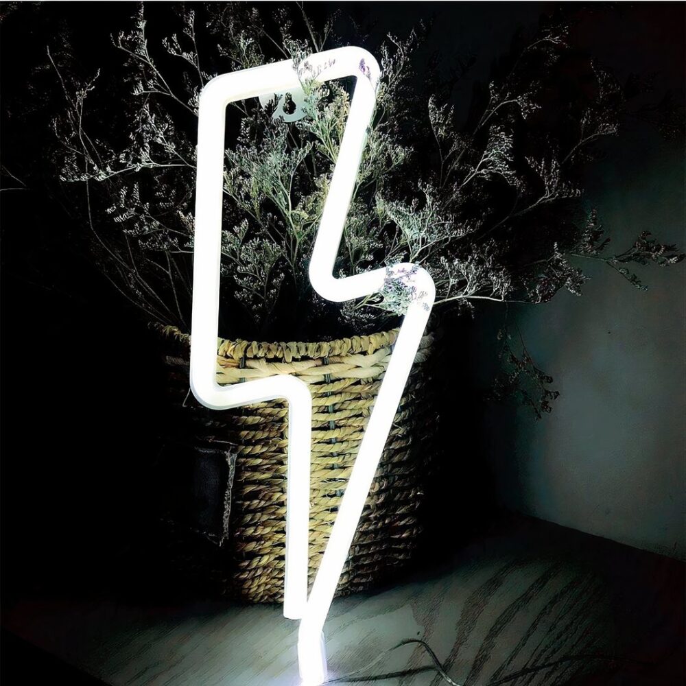 Aplique LED de neón en forma de destello de luz blanca, colocado sobre una mesa y contra una planta.