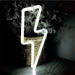 Aplique LED de neón en forma de destello de luz blanca, colocado sobre una mesa y contra una planta.