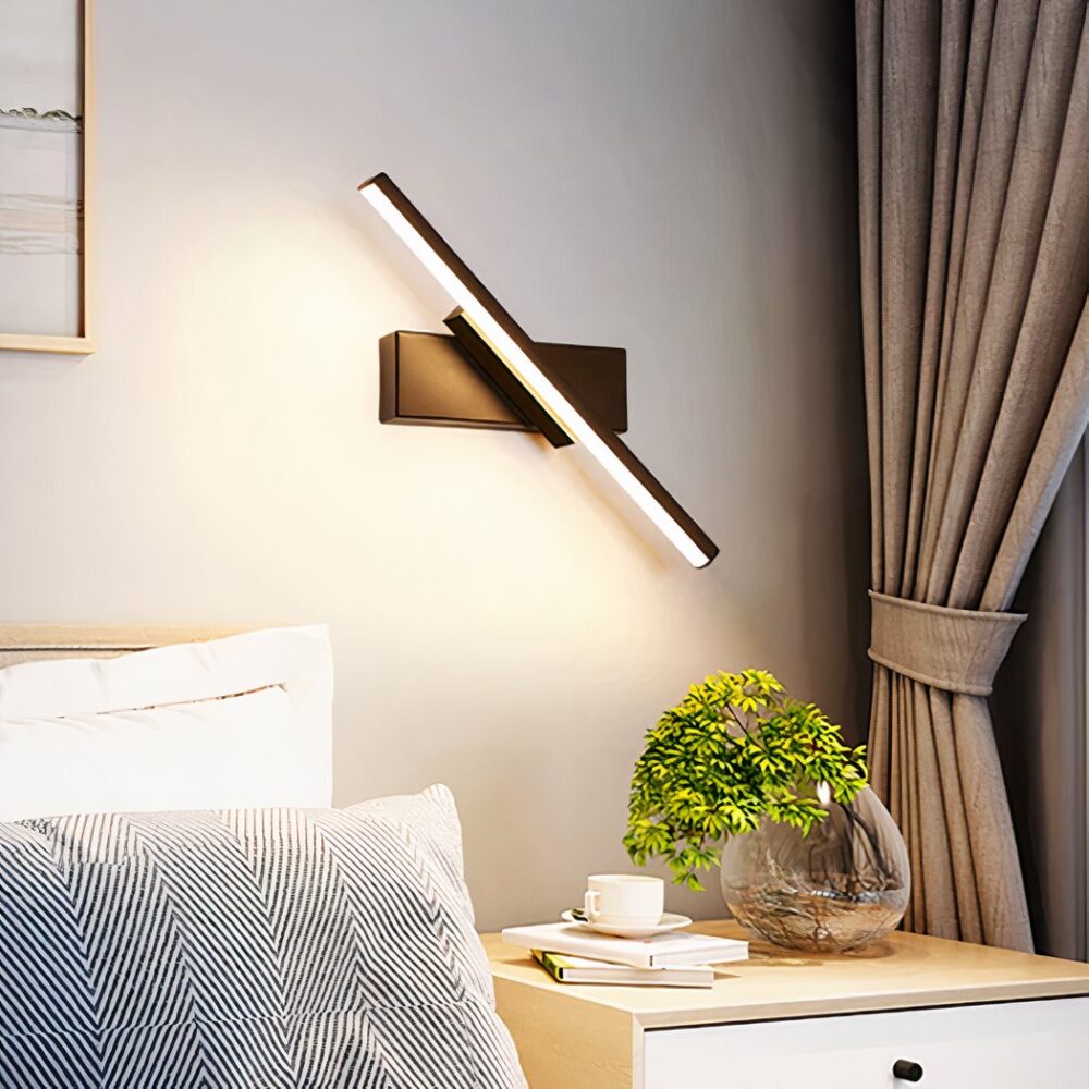 Aplique giratorio de neón negro con luz natural colgado en una pared beige sobre una mesilla de noche con una planta en un jarrón, una pila de libros y una taza de café, junto a una cama con almohadas blancas y grises