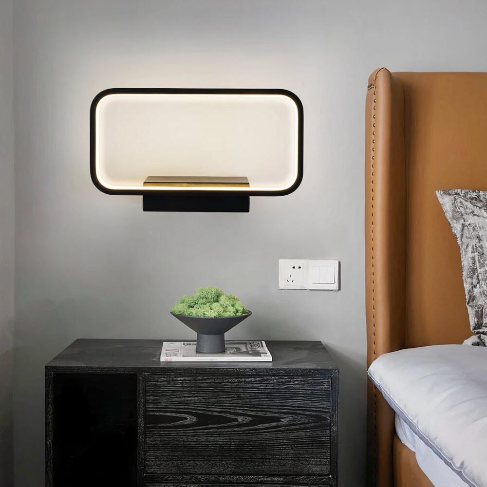Aplique en forma de marco negro luminoso con un soporte rectangular fijado a una pared gris. Una mesilla de noche gris oscuro con una pequeña planta verde debajo. Una cama marrón a la derecha.