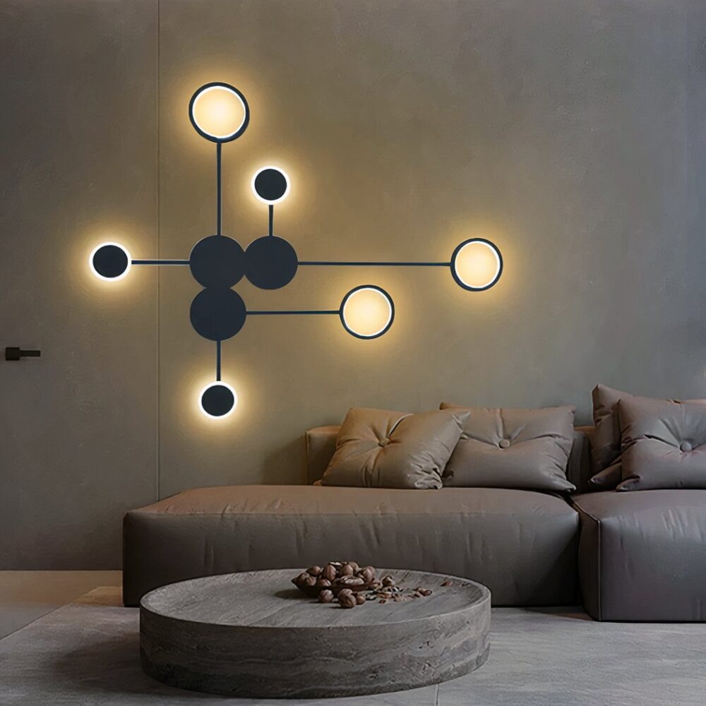 Aplique negro formado por círculos unidos por líneas. 3 círculos negros en el centro y 6 círculos alrededor del aplique. Montado en una pared marrón sobre un sofá marrón con cojines a juego. Mesa de centro redonda marrón y alfombra beige debajo.