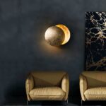 Aplique redondo en 2 partes, una encima de la otra, dorado, con la iluminación entre las 2 partes formando la forma de una luna. Montado en una pared oscura encima de 2 sillones beige y a la izquierda de un marco negro y dorado.