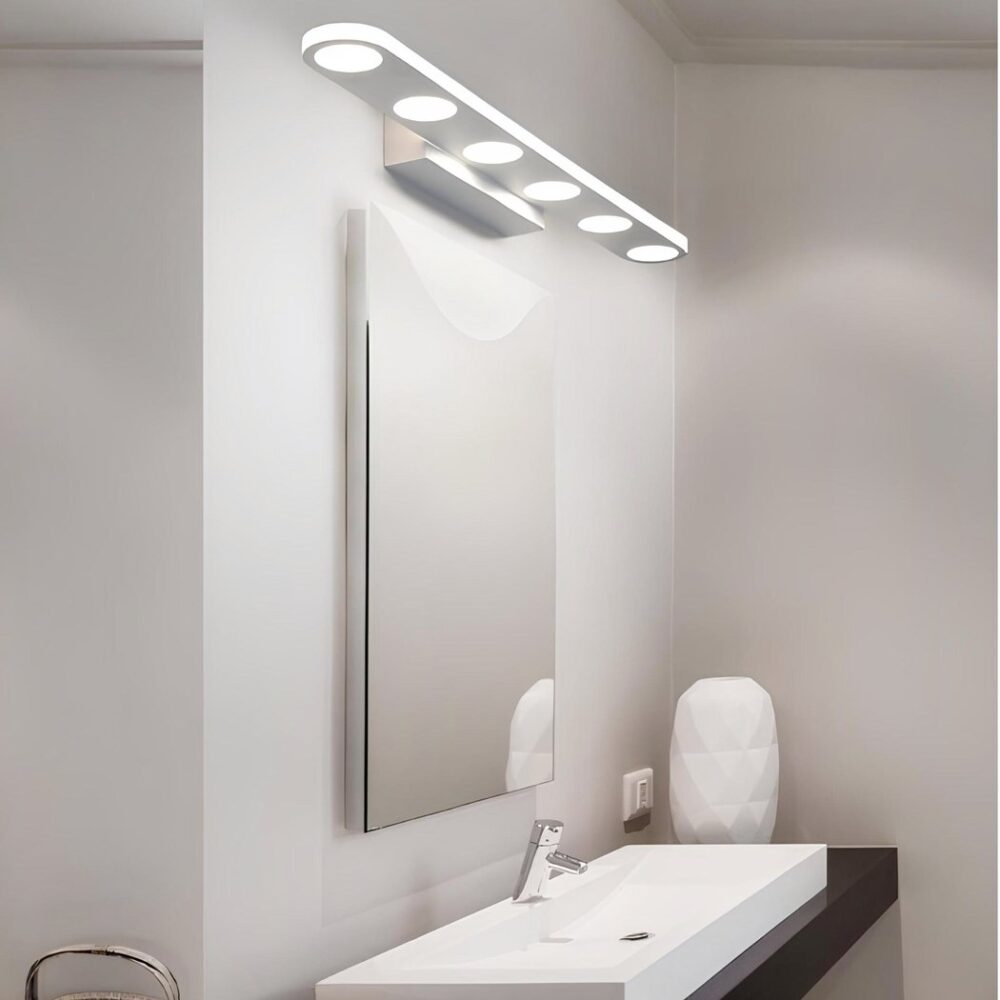 Aplique largo blanco con 6 luces redondas en la parte inferior. Montado en una pared blanca encima de un espejo cuadrado, que a su vez está encima de un lavabo rectangular blanco. El mueble sobre el que se asienta es de color marrón oscuro. En primer plano hay toallas enrolladas con una cesta beige a la izquierda. Al fondo, a la derecha, hay una escultura blanca ovalada.