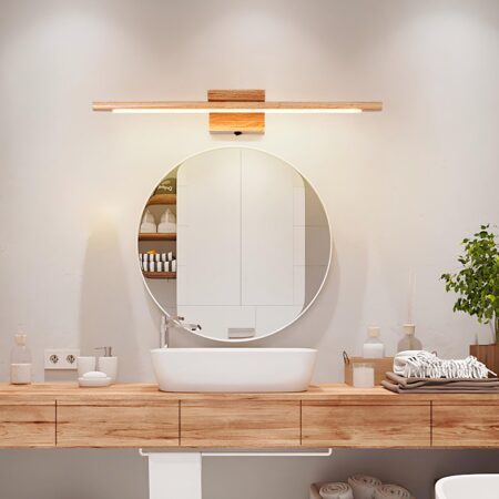 Aplique largo de madera iluminado sobre un espejo redondo. El espejo está encima de un lavabo blanco con encimera de madera con botellas a izquierda y derecha del lavabo, toallas beige a la derecha y una planta verde.