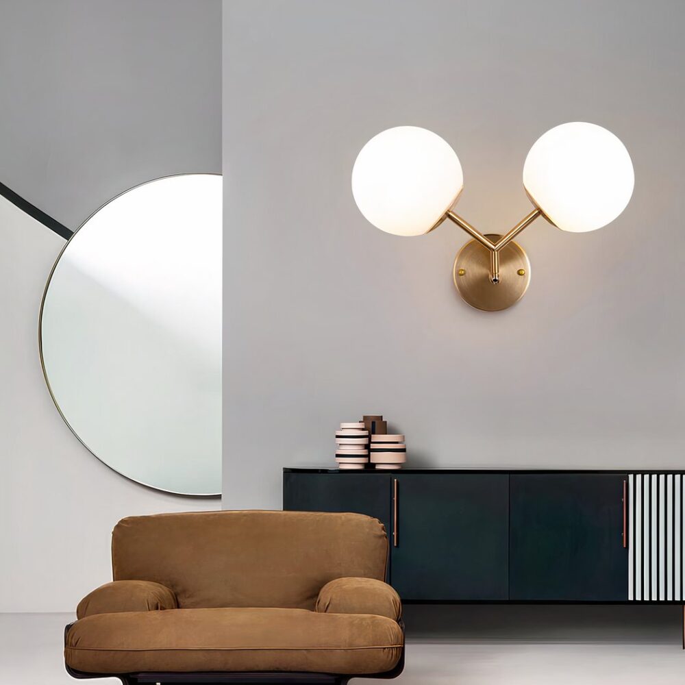 Aplique dorado con 2 bolas luminosas sujetas por una varilla dorada en forma de V. Montado en una pared gris sobre un mueble bajo negro Delante hay un pequeño sillón marrón. A la izquierda, al fondo, hay un gran espejo redondo.