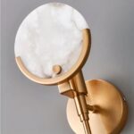 Primer plano de un aplique de pared. Soporte redondo dorado fijado a una pared gris que sostiene un tallo dorado sobre el que se coloca un círculo de mármol. La parte inferior del círculo está sujeta por un borde metálico dorado.