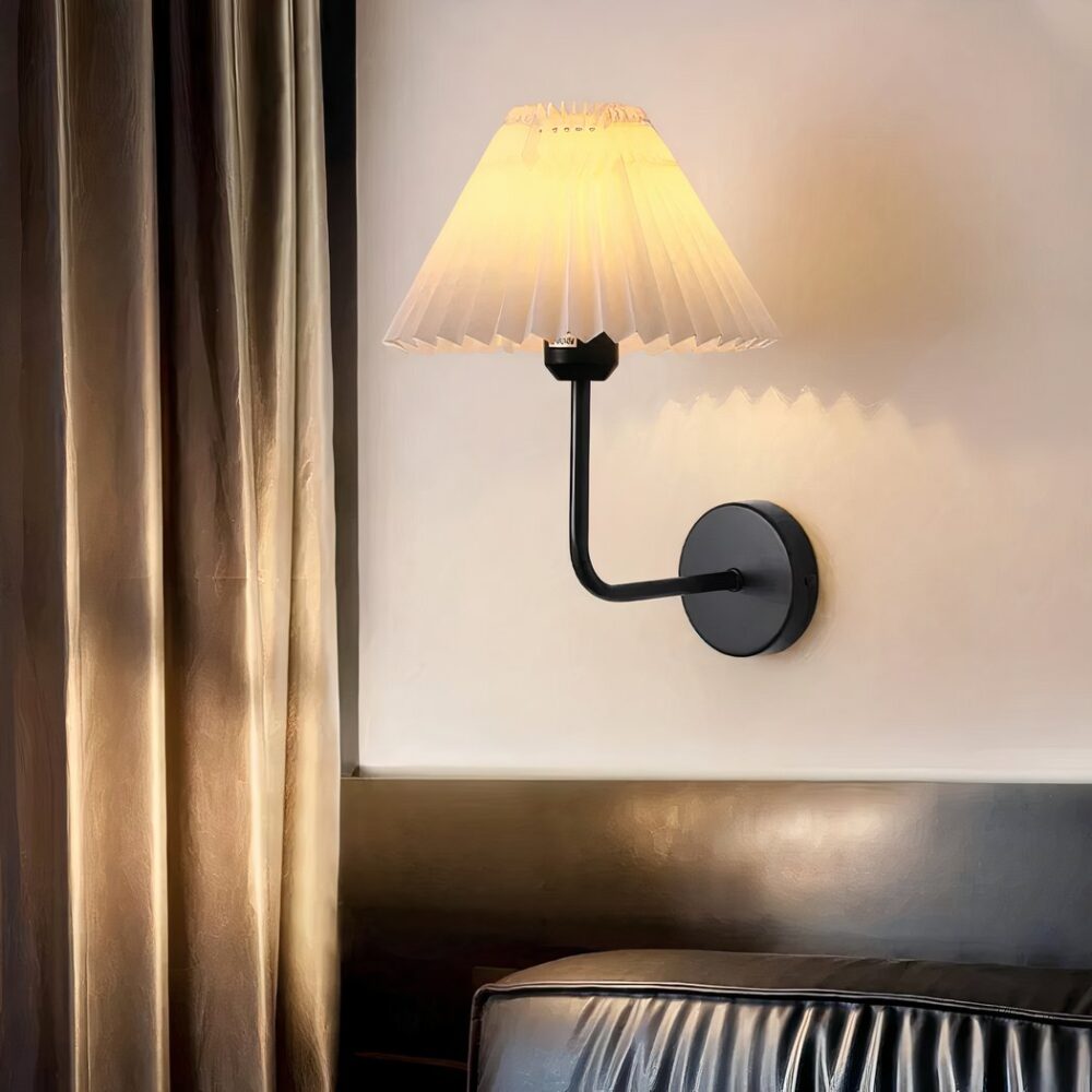 Aplique vintage de salón con pantalla de tela doblada en forma de cono. Está sujeto por una varilla doblada de metal negro sobre un soporte redondo negro fijado a una pared beige. A la izquierda hay una gran cortina marrón, debajo una sección de pared marrón con un gran cojín de cuero marrón delante.