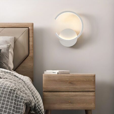 Aplique de aluminio blanco con forma de círculo abierto a la izquierda, como una C invertida. La luz se difunde mediante LED a lo largo de la forma de C. La luz se difunde mediante LED a lo largo de la C. Montado en una pared blanca encima de una mesilla de noche marrón de 2 cajones y a la izquierda de una cama marrón con ropa de cama a cuadros marrones y beige.