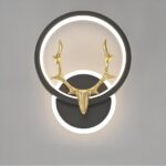 aplique negro, el soporte es redondo con un círculo de luz a su alrededor. Sostiene un círculo de luz en un círculo negro sobre el que se sitúa una cabeza de ciervo dorada. Sobre un fondo gris.