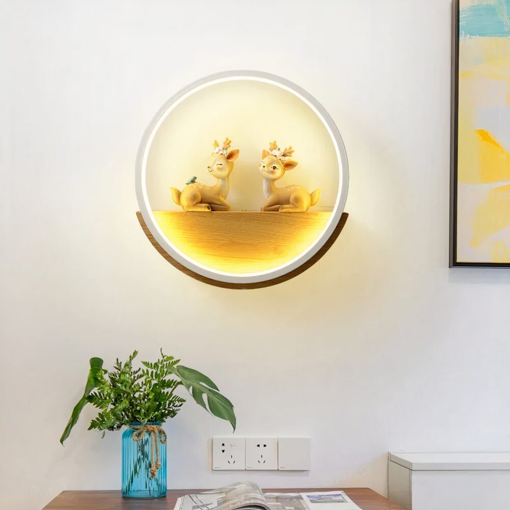 Aplique con forma de círculo blanco. En el interior, hay un soporte de madera en forma de media luna con 2 ciervos enfrentados. El aplique está iluminado y fijado a una pared blanca, con un marco de color a la derecha y debajo un mueble marrón con un jarrón azul y follaje en su interior y una revista.
