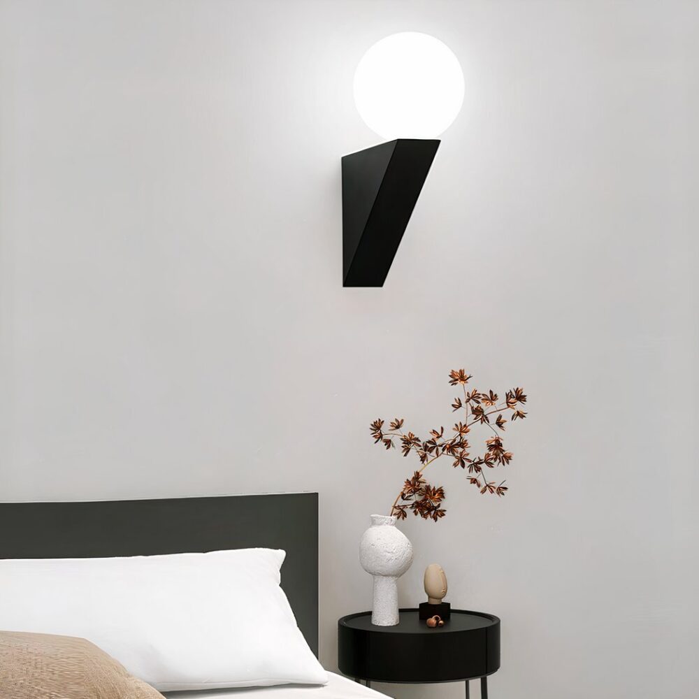 Aplique con forma de soporte negro contra la pared y una bola luminosa en la parte superior. Fijo encima de una mesita de noche negra con un tallo con flores rosas, a la izquierda, una cama con cabecero negro y cojín blanco.