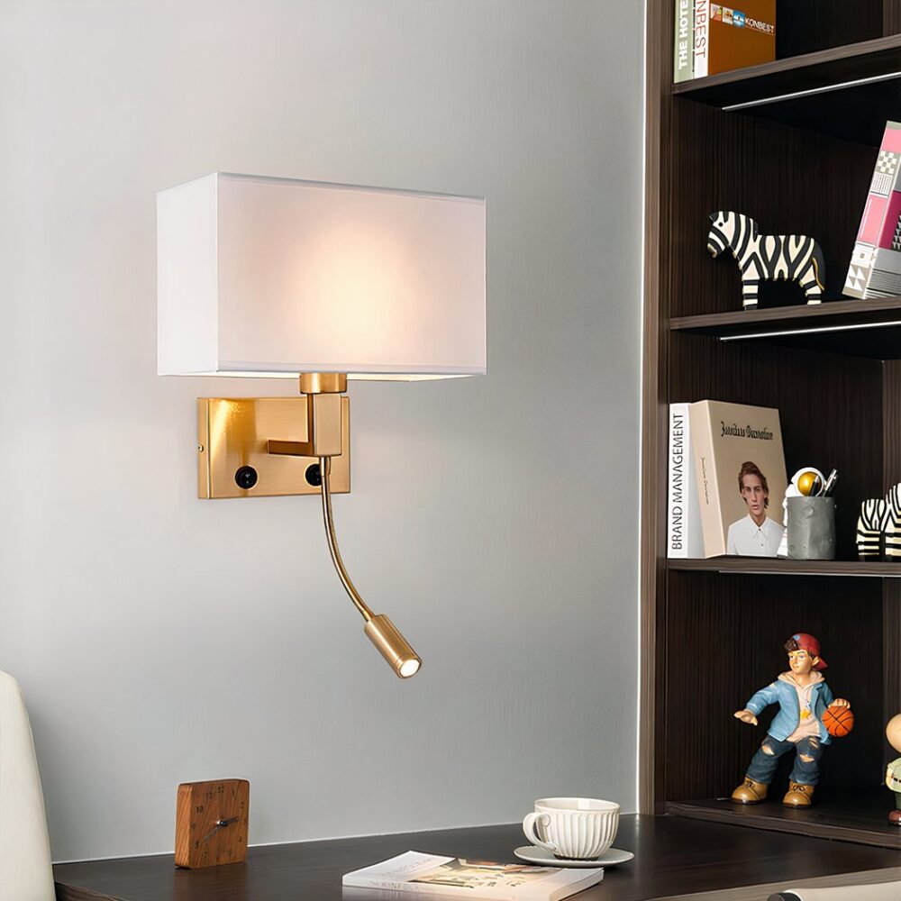 Aplique de pared con base rectangular dorada y tija rectangular curvada hacia arriba. Pantalla de tela blanca. La bombilla está encendida. En la base, dos interruptores negros y una lámpara de lectura flexible dorada. El aplique está fijado encima de un escritorio marrón y a la izquierda de una estantería marrón que contiene libros y figuritas.