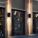 Fachada de tienda con escaparates enmarcados por pilares de hormigón en cada uno de los cuales se ha fijado un aplique. El aplique es rectangular y de color negro, y difunde un haz de luz cálida por encima y por debajo.