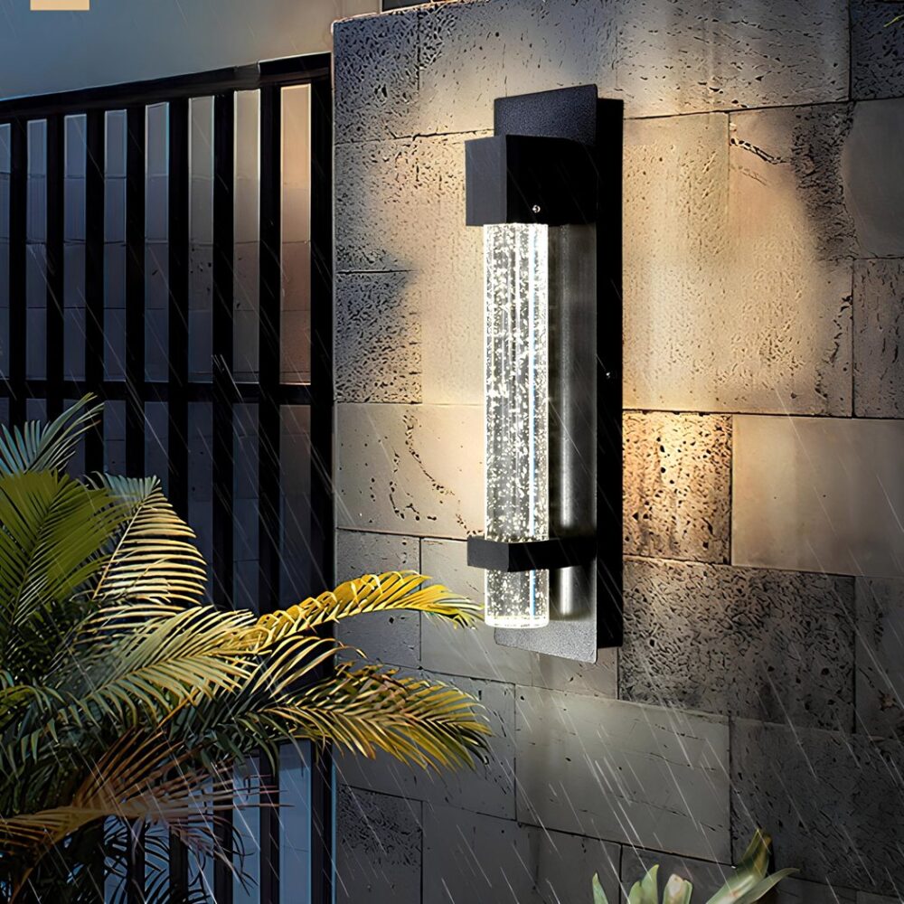 Aplique de exterior en forma de tubo de burbuja en una estructura de aluminio negro fijada a un muro de piedra. A la izquierda se ve una gran verja negra, parte de una palmera delante del muro y lluvia cayendo.