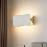 Aplique moderno rectangular de metal blanco con tapa basculante para dirigir la luz. Cortina marrón a la izquierda. Montado en una pared blanca.