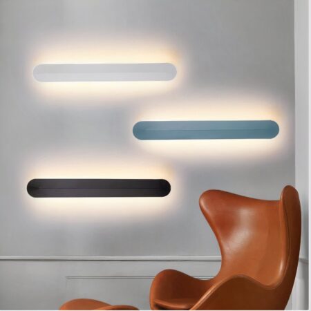 Aplique moderno minimalista en tres versiones, blanco, negro y azul pato, colgado en una pared junto a un sillón de orejas de cuero coñac.