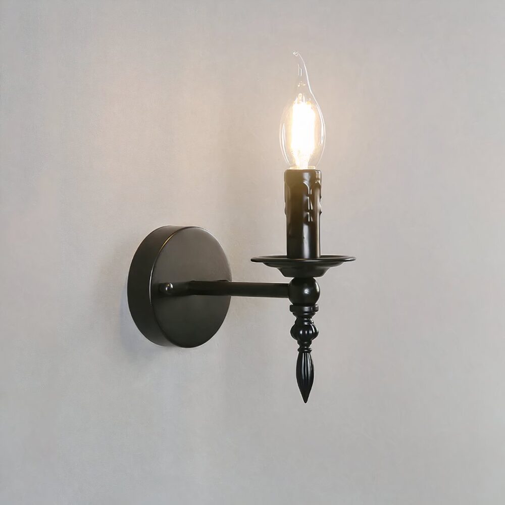 Aplique de metal negro con forma de candelabro. La bombilla encendida tiene forma de llama. Montado en una pared blanca.
