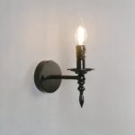 Aplique de metal negro con forma de candelabro. La bombilla encendida tiene forma de llama. Montado en una pared blanca.
