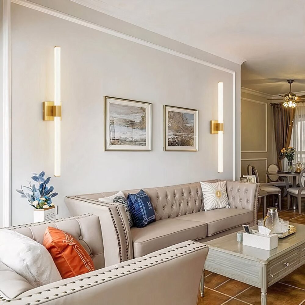 Aplique rectangular de diseño tubular en su versión dorada e instalado en una pared blanca de un salón, a ambos lados de un sofá beige.